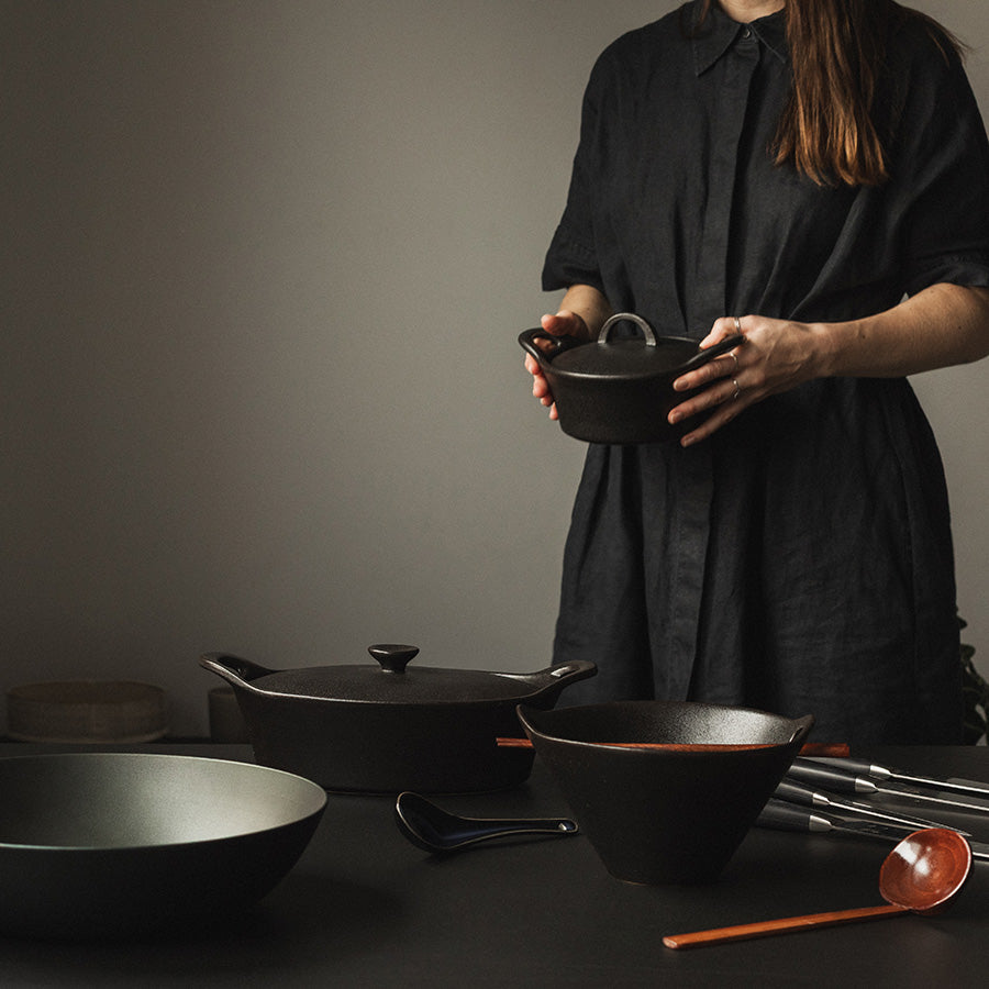 Kvinne som holder en sort ildfast form med lokk, med andre servise og kjøkkenutstyr på bordet.