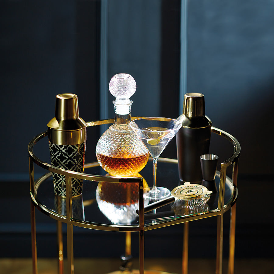 En elegant cocktail på et glassbord med Barcraft cocktailsett i bakgrunnen, inkludert to cocktailshakere og en dekorativ karaffel.