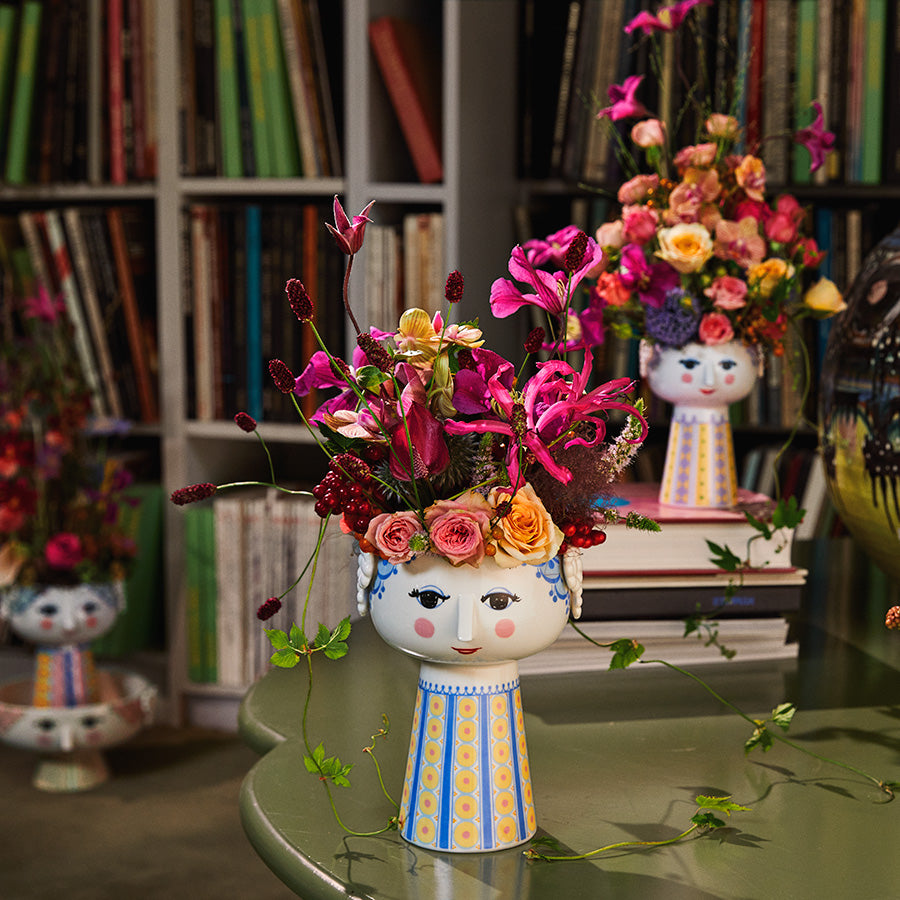 Bjørn Wiinblad Eva vase med blomsterarrangement i rosa og lilla toner, plassert blant bøker, skaper en livlig interiørdetalj.