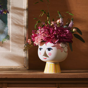 Bjørn Wiinblad potteskjuler med blomsteroppsats, dekorert ansikt og fargerike detaljer, plassert på et bord med kunstverk i bakgrunnen.