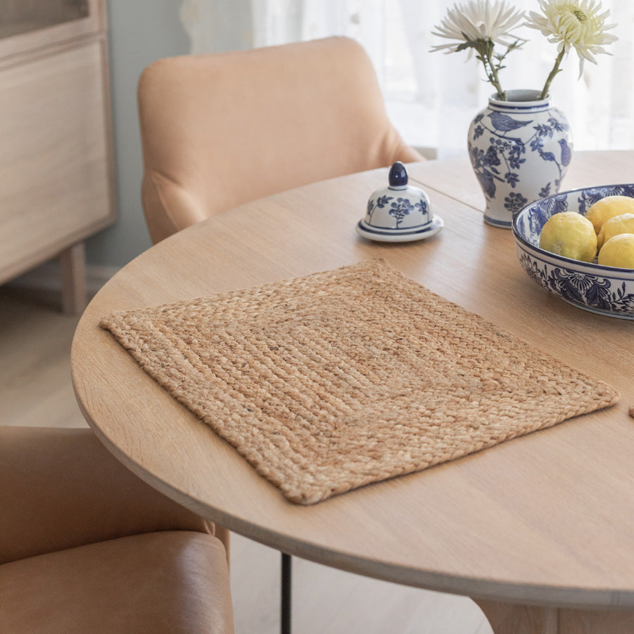 Dekkebrikke i jute på et rundt spisebord, omringet av stoler og dekorative elementer som en vase med blomster og en skål med sitroner.