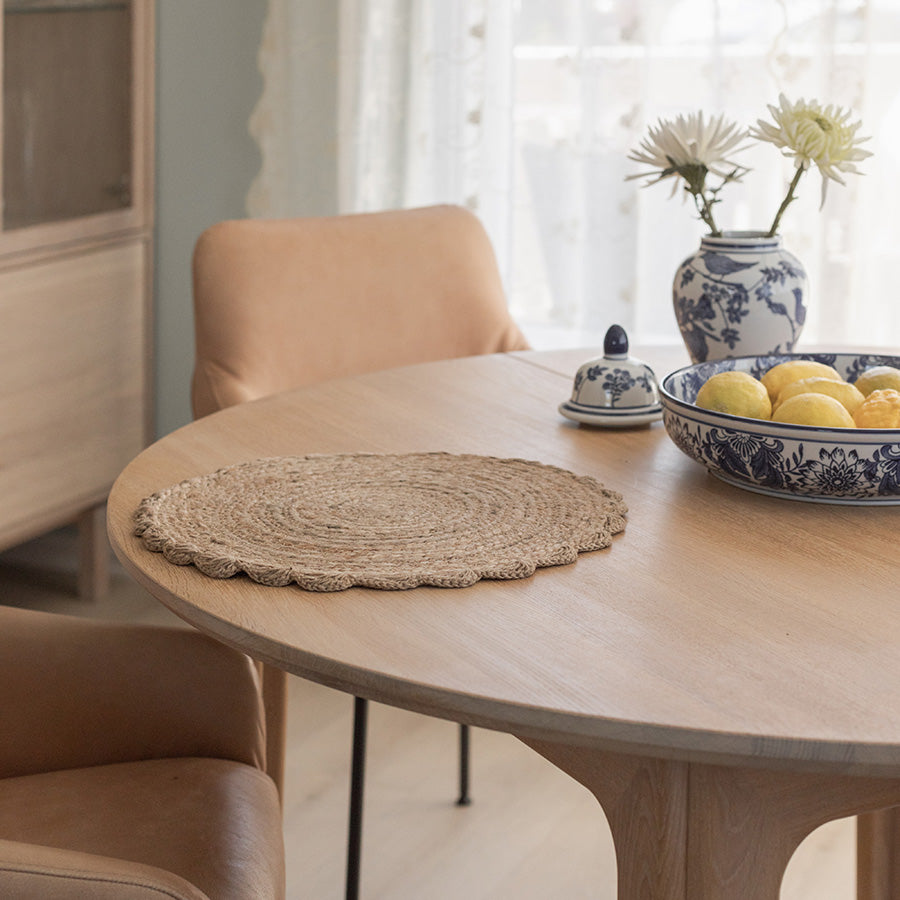 Rund dekkebrikke i jute på et lyst, moderne spisebord med stoler, dekorert med frukt og blomster.