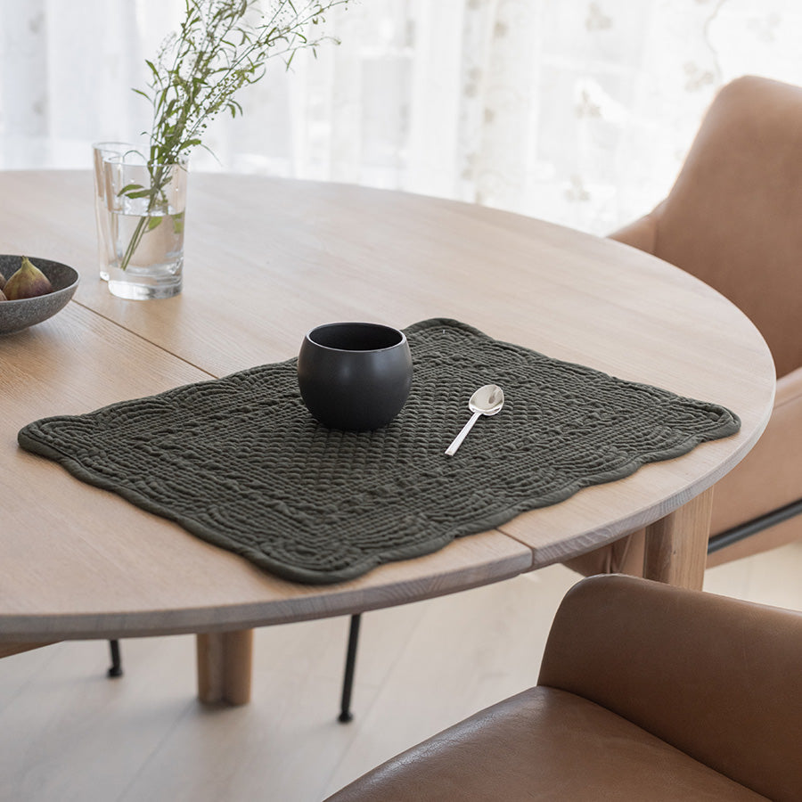 Dekkebrikke i grønn velour på et lyst rundt bord, med en sort kopp og en skje, gir en elegant og praktisk borddekking.