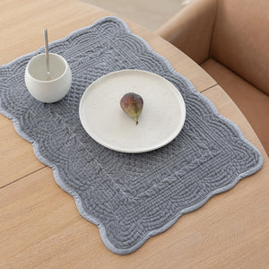 Dekkebrikke i quiltet velour med lys grå farge, elegant plassert på et bord med en tallerken og en frukt. Perfekt for borddekking.