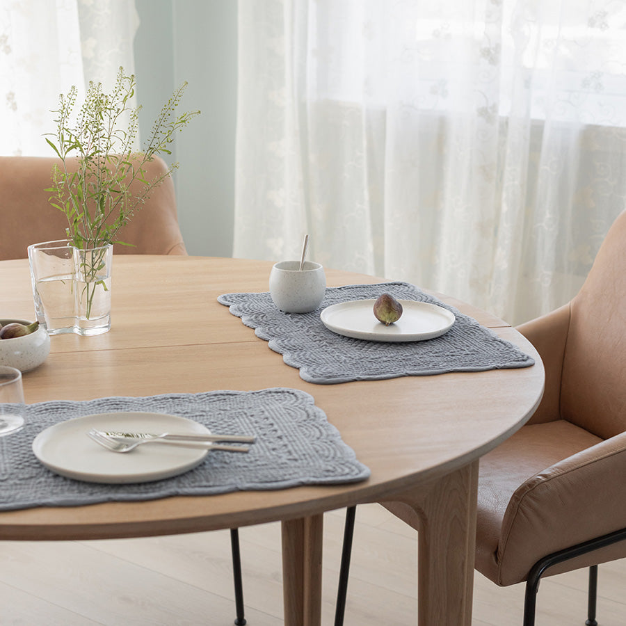 To dekkebrikker i quiltet velour på et rundt bord, sammen med tallerkener og glass, skaper et innbydende og moderne bordmiljø.