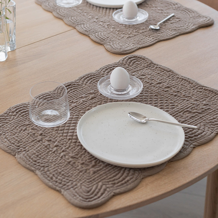Dekkebrikke i sandfarget velour med quiltet mønster på et bord, med en hvit tallerken, glass og egg på en elegant borddekking.