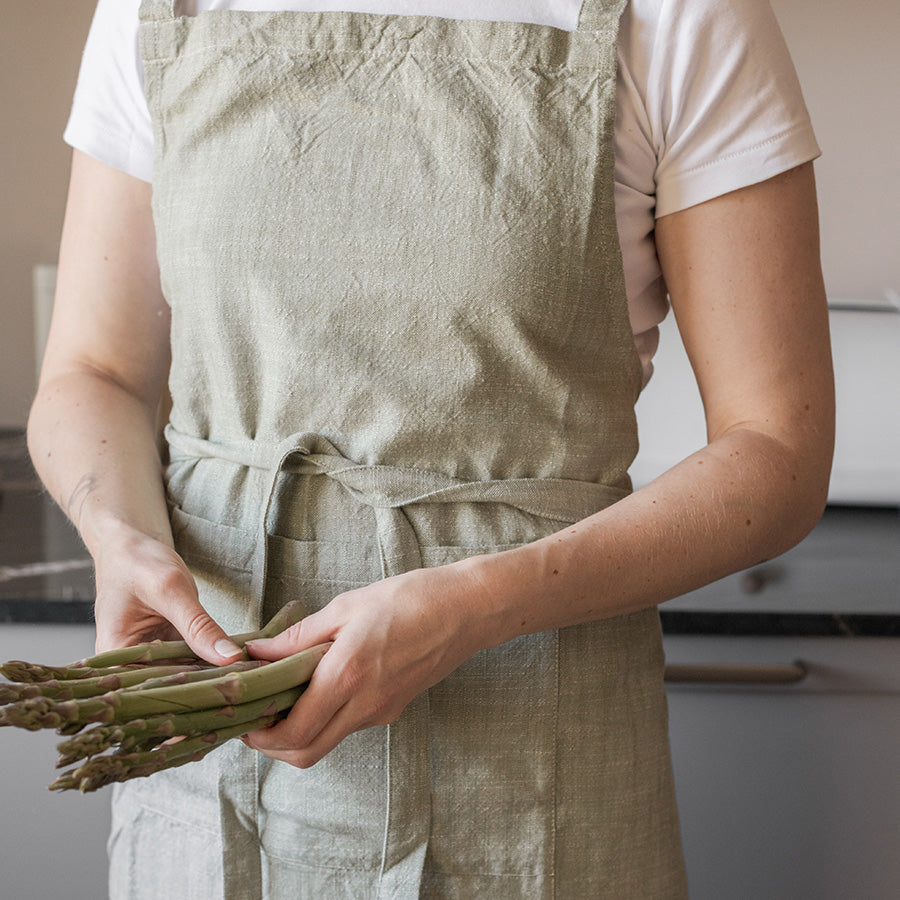 En person iført et grønt kjøkkenforkle i 100% bomull, holder friske asparges. Forkleet har en dempet stil og passer perfekt til både matlaging og grilling.