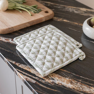 Christiania Glasmagasin gryteklut i lys grønn/hvitt, quiltet design, 20x20 cm, plassert på en kjøkkenbenk med grønnsaker, perfekt for håndtering av varme kjeler.