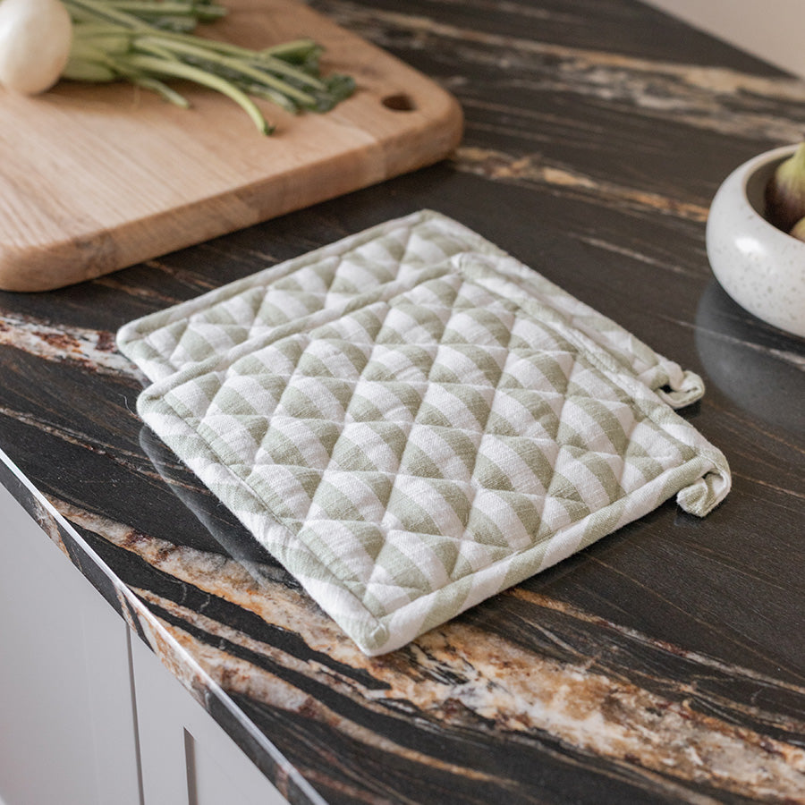 Christiania Glasmagasin gryteklut i lys grønn/hvitt, quiltet design, 20x20 cm, plassert på en kjøkkenbenk med grønnsaker, perfekt for håndtering av varme kjeler.