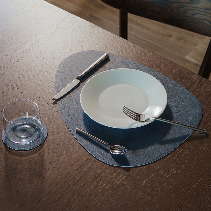 Borddekkingsbilde med mørk blå spisebrikke, tallerken, kniv, gaffel og glass.