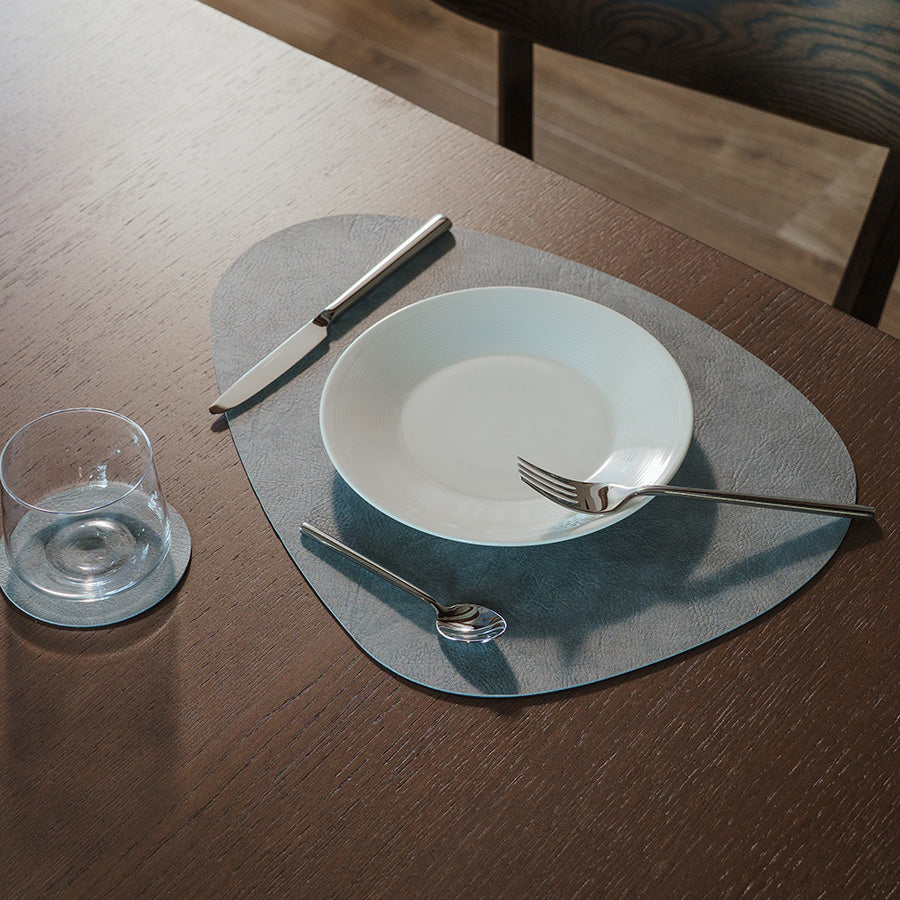 Borddekkingsbilde med grå spisebrikke, tallerken, kniv, gaffel og glass.