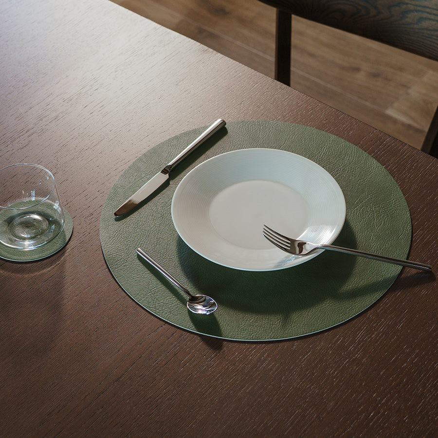 En grønn spisebrikke på et bord med tallerken, bestikk og glass, skaper en koselig borddekkning.