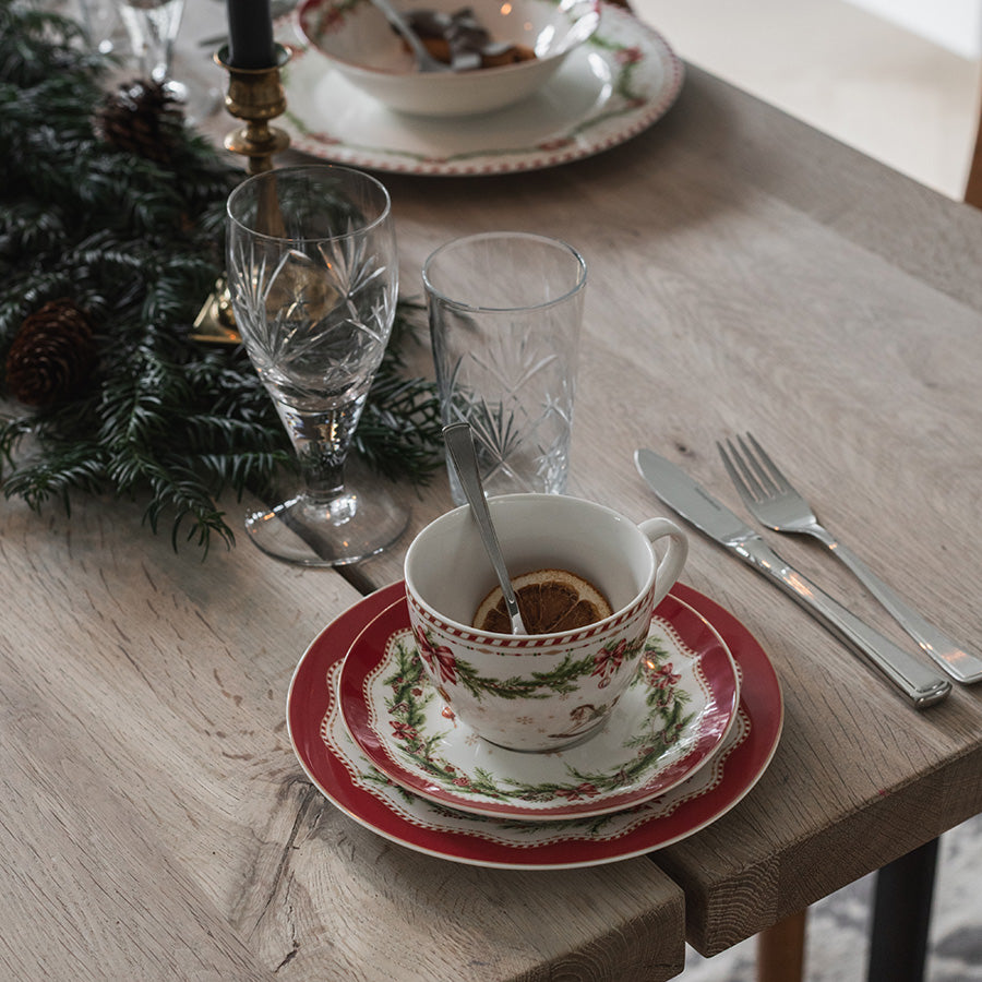 Juleglede kopp og skål med kaffe på juledekket trebord, omgitt av granbar, glass og bestikk