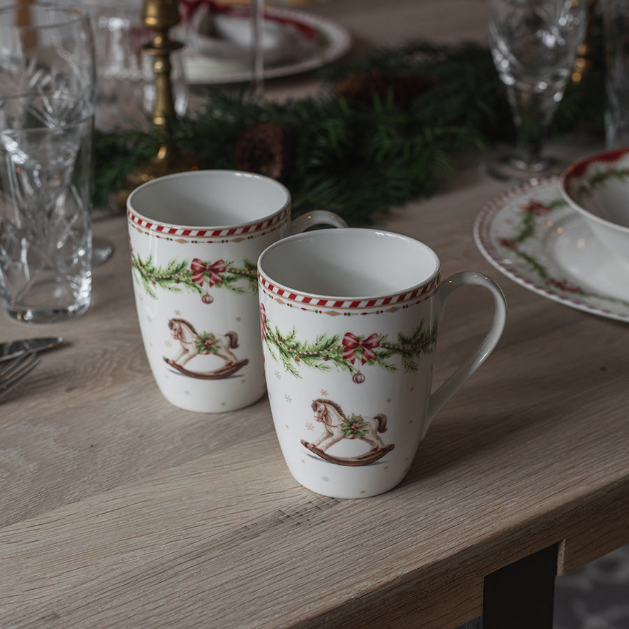 To Juleglede krus med julemotiv og gyngehest, dekorativt plassert på festdekket julebord