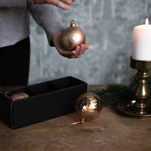 Hånd som holder en dekorert gullfarget julekule i glass ved siden av gaveeske og levende lys