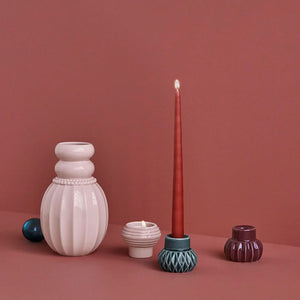 Lysestake kombinert med vasen fra Dottis Nordic Design, med en rød telys og keramiske elementer i mørk, pudder, og aubergine, stilfullt presentert mot en rosa bakgrunn.