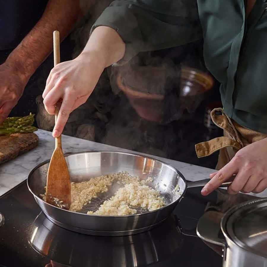 Person som rører i risotto i All Steel Pure stekepanne fra Fiskars Kjøkken, med grønnsaker og en annen person som forbereder ved siden av.
