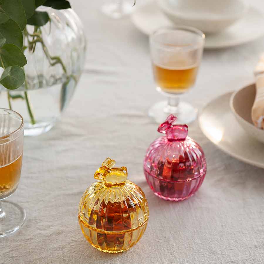 To Hadeland Glassverk glasskåler, en gul og en rosa, dekorert med hare-design, plassert på et dekket bord med drikke og serveringsfat.
