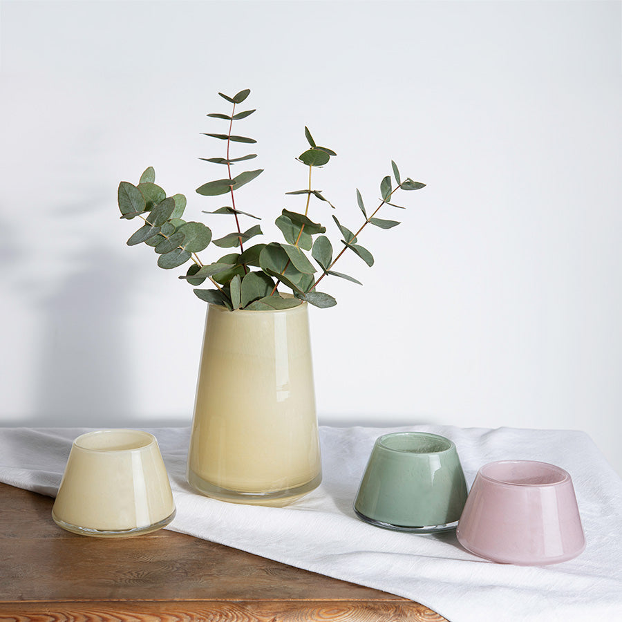 Samling av Hadeland Glassverk lyslykter i ulike farger, inkludert gul, grønn og rosa, med en elegant stil og dekorative blomster i en vase.