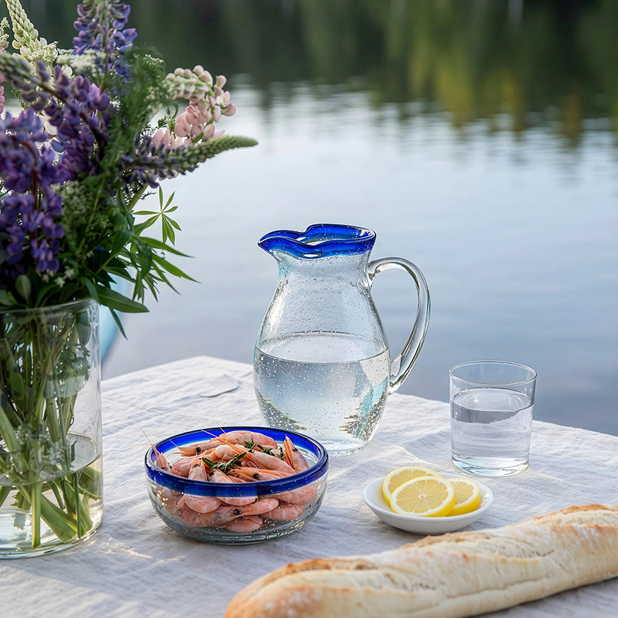 Hadeland Glassverk Skjærgård mugge i naturskjønt miljø, fylt med vann og sitron, omringet av sjømat og blomster.
