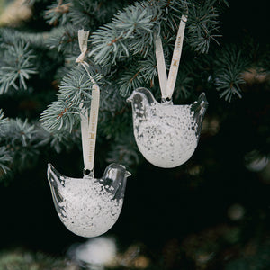 To hvite, spettet glassornamenter formet som turtelduer henger sammen på en grangren