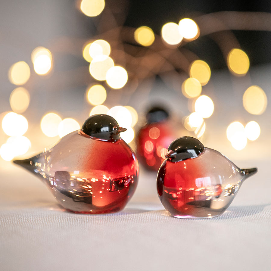 Dompap glassfigur fra Hadeland Glassverk, med rød og svart farge, står på et bord med gyldne lys i bakgrunnen.