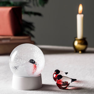 Dompap glassfigur fra Hadeland Glassverk ved siden av en snøkule med dompap motiv, plassert på et elegant dekket bord.