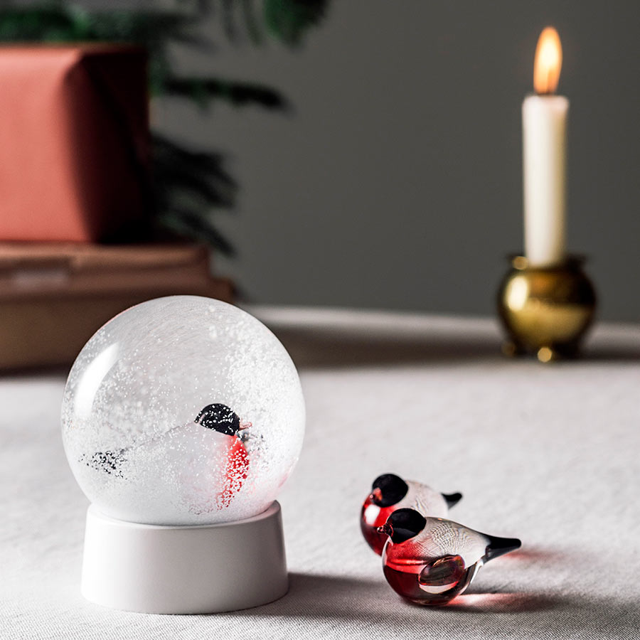 Dompap glassfigur fra Hadeland Glassverk ved siden av en snøkule med dompap motiv, plassert på et elegant dekket bord.