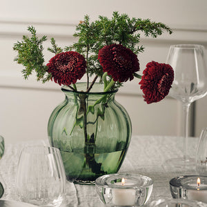 Grønn Hadeland Glassverk Ella vase på 22 cm med mørkerøde blomster, omgitt av glass og tente lys på et elegant spisebord