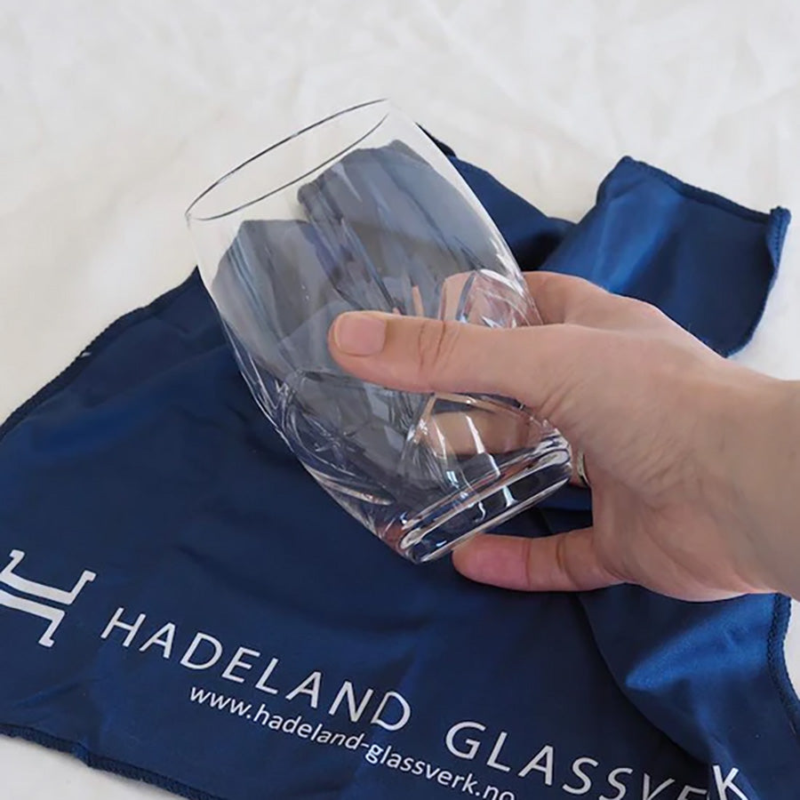 Hånd som holder et glass med Hadeland Glassverk glassklut i blått, demonstrerer effektiv rengjøring og polering.