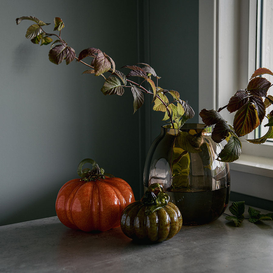 To stilige gresskar i glass fra Hadeland Glassverk plassert på et bord med en vase, omfavnet av høstfarger og stilige grener, ideelt for høstinnredning.