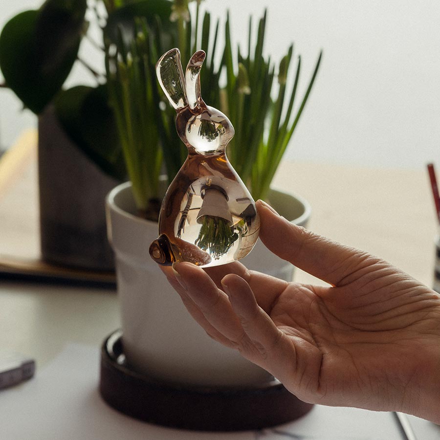 Close-up of a hand holding a glistening glass Hare figurine from Hadeland Glassverk, surrounded by green plants.