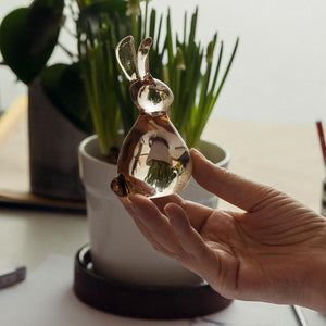 Close-up of a hand holding a glistening glass Hare figurine from Hadeland Glassverk, surrounded by green plants.