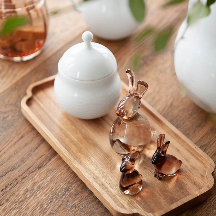 A wooden tray displaying multiple glass Hare figurines from Hadeland Glassverk, along with a white sugar bowl, set in a cozy setting.