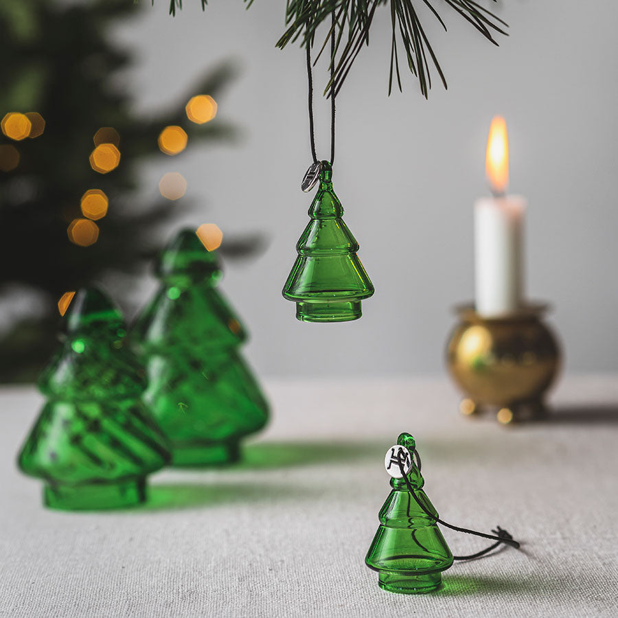 Hadeland Glassverk juletrepynt, flere glassgrantrær i vintergrønn farge hengt fra en gren med telys i bakgrunnen.