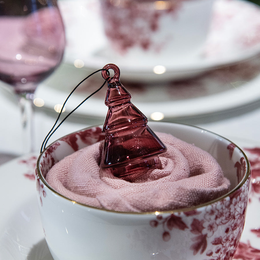 En rosa gran hengepynt fra Hadeland Glassverk plassert i en kopp med serviett, ideell for borddekorasjon og gaveinnpakning.