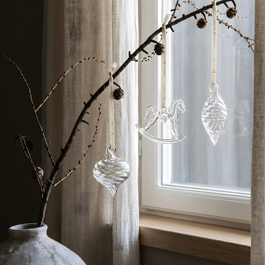Klare glassheng fra Hadeland Glassverk dekorert på en gren foran et vindu med naturlig dagslys