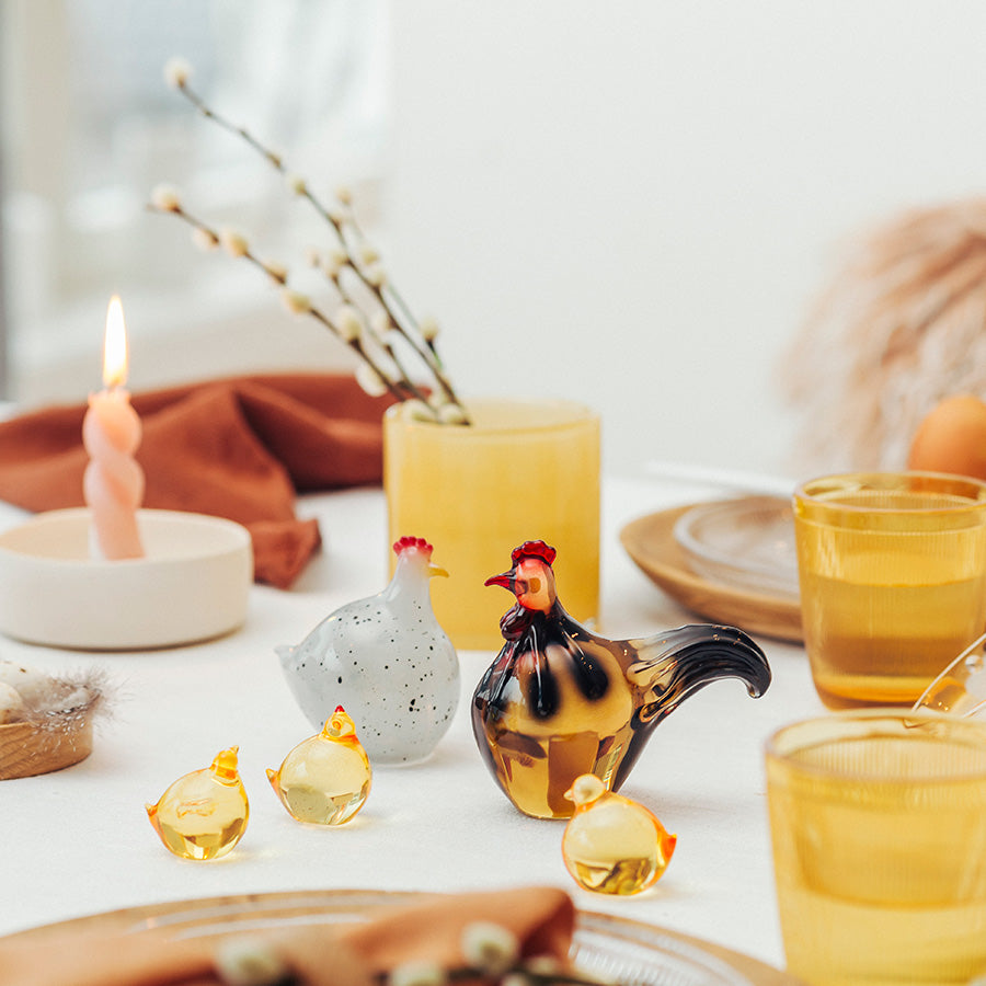 Stilig påskepynt i en feiring med Hadeland Glassverk figurene, en hane, en høne og glasskyllinger plassert på et bord med stearinlys.