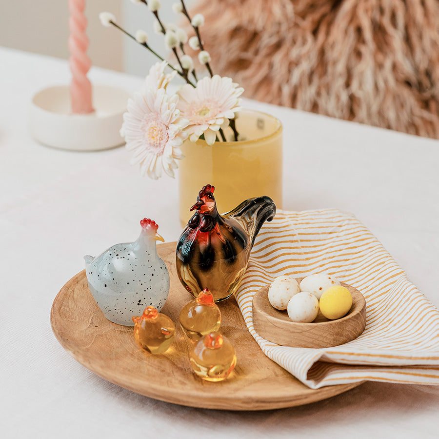 Sjarmerende påskepynt fra Hadeland Glassverk med hane, høne og glasskyllinger, presentert på et trefat med påskeegg.