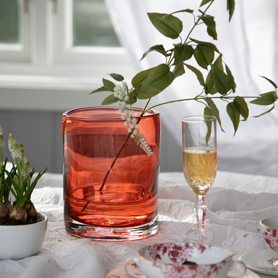 Hadeland Glassverk tulipanvase med en elegant rød tone, fylt med grønne grener og nærliggende glass med drikke, på et bord.