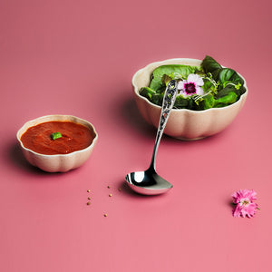 Hardanger Bestikk Kristin sauseøse brukt til servering av saus og salat, sammen med pyntede skåler på en rosa bakgrunn.