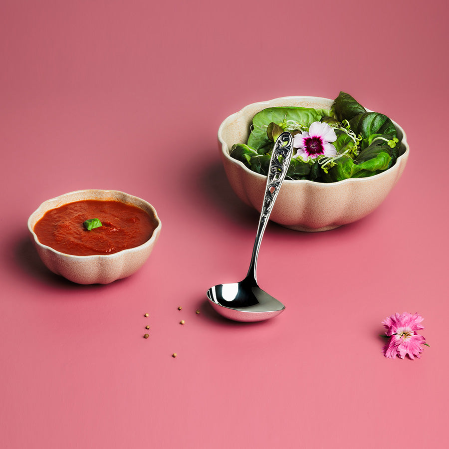 Hardanger Bestikk Kristin sauseøse brukt til servering av saus og salat, sammen med pyntede skåler på en rosa bakgrunn.