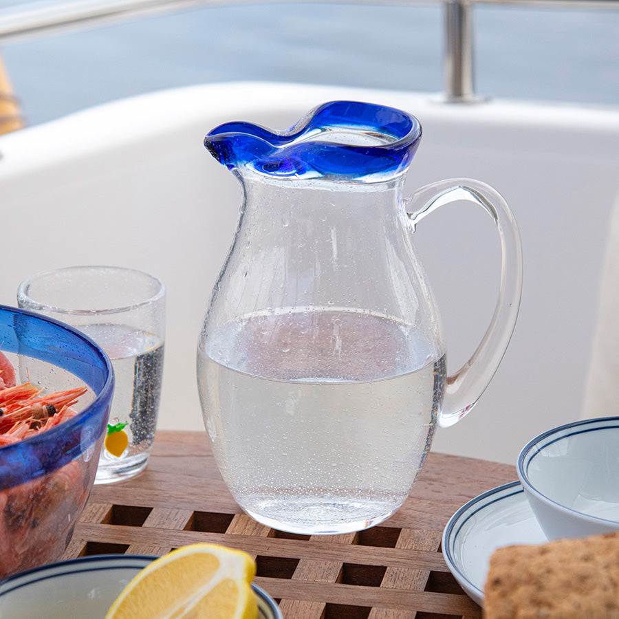 Mugge fra Hadeland Glassverk fylt med vann, plassert på et bord med mat, ideell for sommerbesøk.