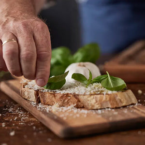 Hånd som strør ferske urter over en skive brød, plassert på et serveringsbrett, som viser brettets praktiske bruk til matservering.