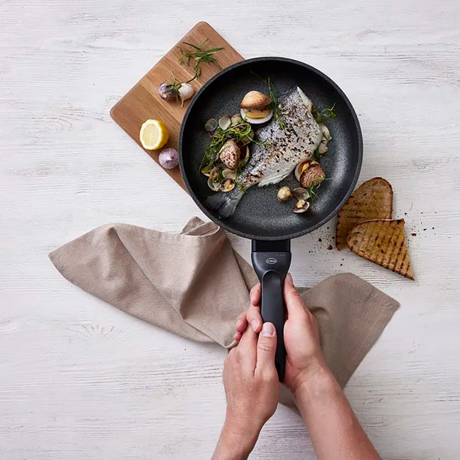 Kokk holder en stekepanne med fisk og grønnsaker, med et serveringsbrett i bakgrunnen, illustrerer allsidigheten til brettet i matlaging og servering.