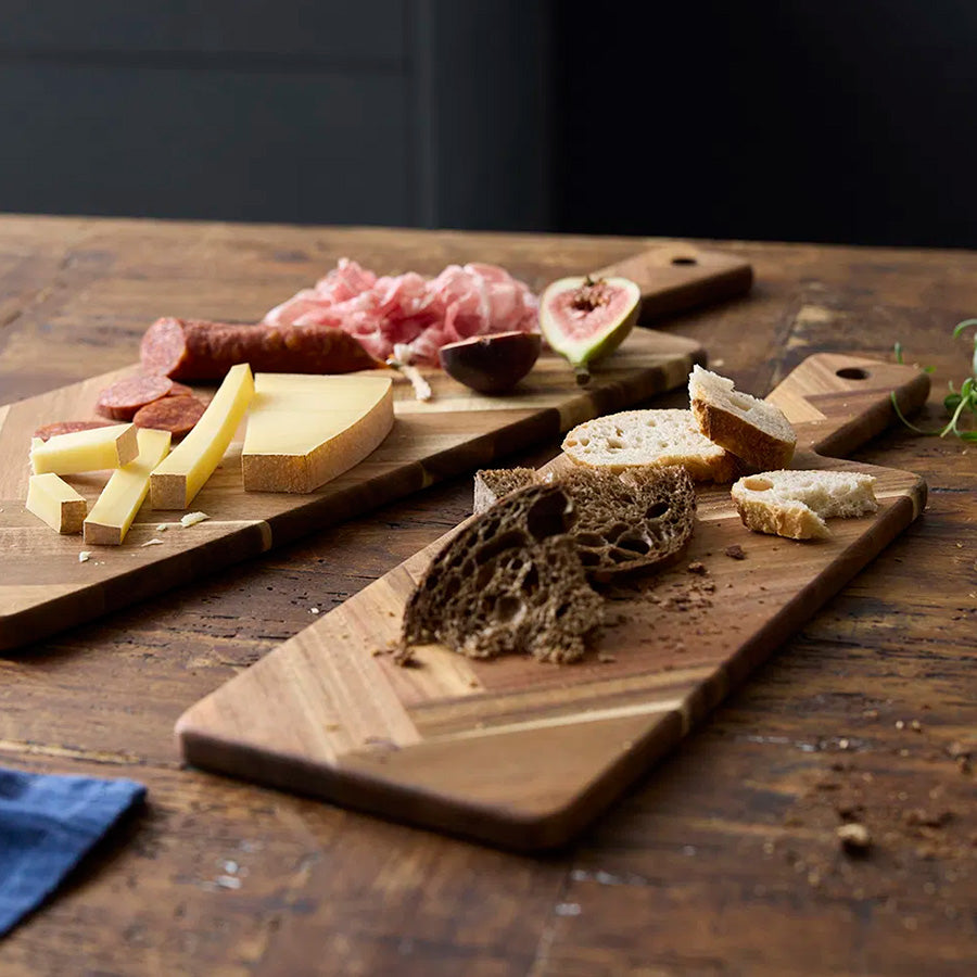 Holm Serveringsbrett Fiksebensmønster 55x20cm Akasie, trefjøl med assorterte oster og spekemat på en rustikk bordflate.