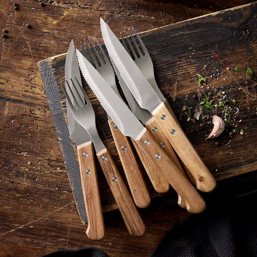 Nærbilde av Holm biffbestikk blant urter og hvitløk, inkludert både gafler og kniver, på en rustikk treplate, passende for gourmetmiddag.