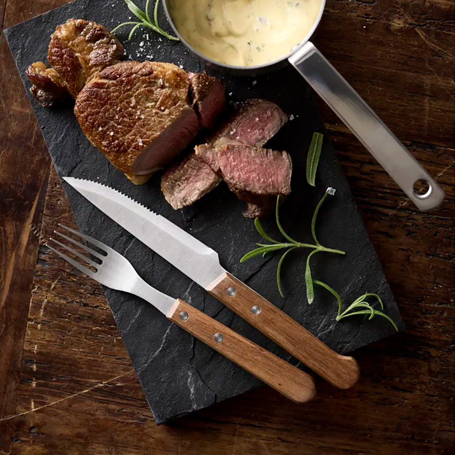 Holm biffbestikk satt opp ved siden av en saftig biff på en steinplate, med en skål med saus i bakgrunnen, ideelt for måltider med grillmat.