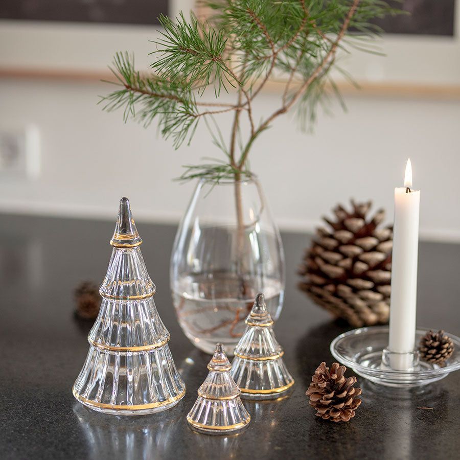 Holmegaard Fairytales juletre i klart glass med gulldetaljer, i en vinterdekorasjon med kongler og små trær på et kjøkkenbord.
