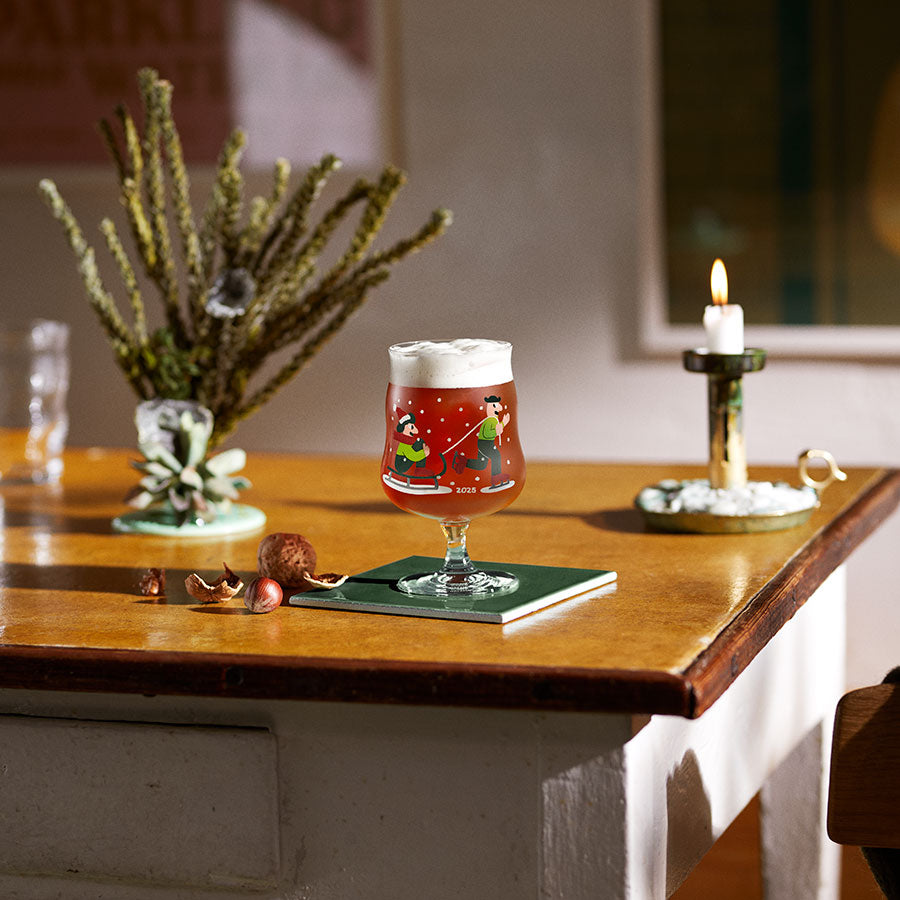 Holmegaard ølglass på bord med dekorativt julemotiv, fylt med øl og pisket krem, skaper en koselig atmosfære.